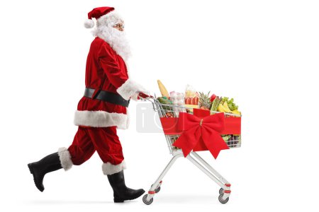 Photo for Santa Claus running and pushing a shopping cart with food tied with a red bow isolated on white background - Royalty Free Image