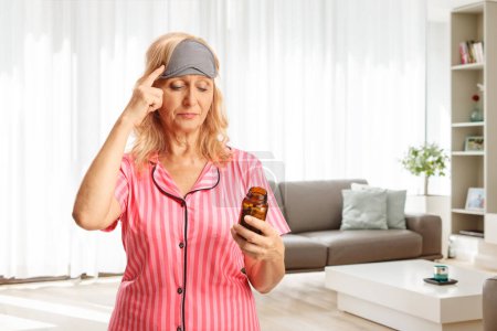 Téléchargez les photos : Femme en pyjama tenant une bouteille de pilules et tenant sa tête à la maison dans un salon - en image libre de droit