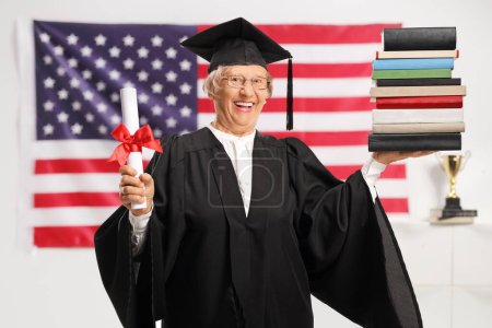 Photo for Senior woman in a graduation gown holding a diploma and a pile of books with the USA flag in the background - Royalty Free Image