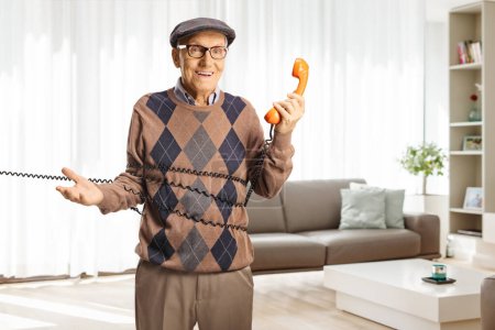 Photo for Elderly man tangled in a cable from a vintage rotary phone standing in a living room and smiling - Royalty Free Image