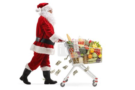 Photo for Full length profile shot of Santa Claus walking and pushing a shopping cart with food and money falling isolated on white background - Royalty Free Image