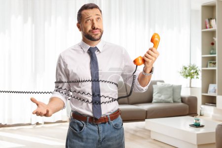 Photo for Irritated young man tied with a rotary phone cable at home in a living room - Royalty Free Image