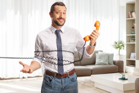 Photo for Bored young man standing tangled in a cable from a vintage rotary phone in a living room - Royalty Free Image
