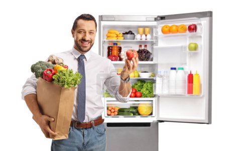 Photo for Cheerful young man holding a grocery bag and a tomato in front of a fridge isolated on white background - Royalty Free Image