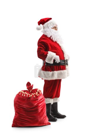 Foto de Perfil completo plano de Santa Claus de pie junto a su saco en el suelo aislado sobre fondo blanco - Imagen libre de derechos