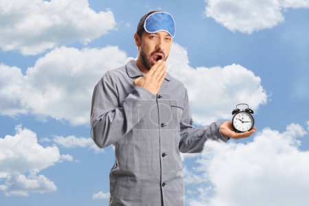 Téléchargez les photos : Jeune homme en pyjama avec un masque de sommeil tenant un réveil et bâillant avec des nuages et du ciel en arrière-plan - en image libre de droit