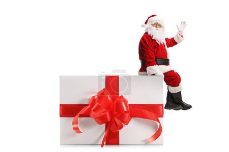 Foto de Santa Claus sentado en una caja de regalo y saludando aislado sobre fondo blanco - Imagen libre de derechos