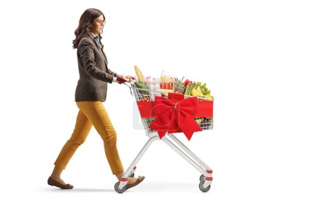Photo for Full length profile shot of a young woman walking with a shopping cart tied with a red ribbon isolated on white background - Royalty Free Image