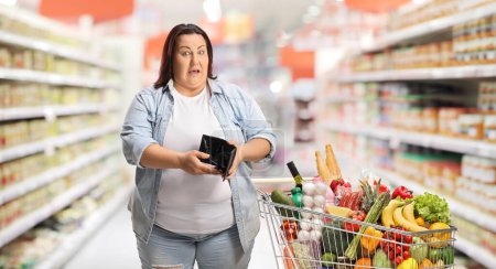Foto de Mujer corpulenta con un carrito de compras sosteniendo una cartera vacía en un supermercado - Imagen libre de derechos