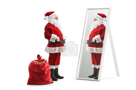 Téléchargez les photos : Photo de profil pleine longueur du père Noël debout à côté de son sac et regardant un miroir isolé sur fond blanc - en image libre de droit
