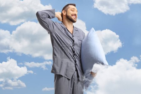 Photo for Young man in pajamas holding a pillow and stretching with blue sky and clouds in the background - Royalty Free Image