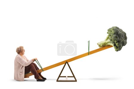 Photo for Full length profile shot of an elderly woman and a broccoli on a seesaw isolated on white background - Royalty Free Image