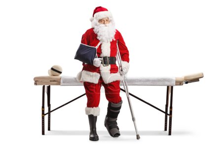 Photo for Sad santa claus with injured leg and arm sitting on a mat table isolated on white background - Royalty Free Image