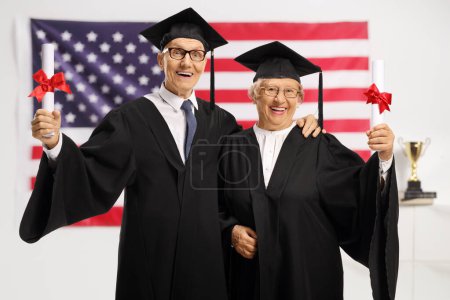 Photo for Senior man and woman in graduation gowns holding diplomas and smiling in front of a USA flag - Royalty Free Image