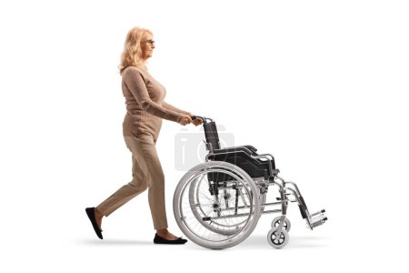 Téléchargez les photos : Photo de profil pleine longueur d'une femme mature marchant et poussant un fauteuil roulant vide isolé sur fond blanc - en image libre de droit