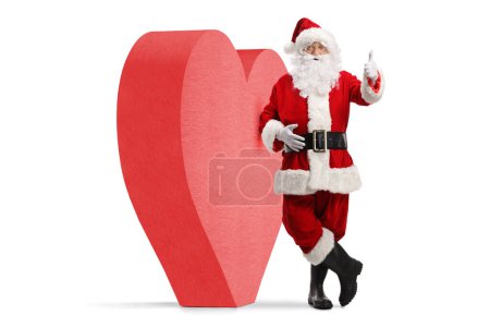 Téléchargez les photos : Portrait complet du Père Noël appuyé sur un cœur rouge et les pouces gestuels vers le haut isolé sur fond blanc - en image libre de droit