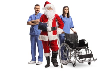 Foto de Santa Claus lesionado con un soporte para los pies y una honda para los brazos de pie con un equipo médico aislado sobre fondo blanco - Imagen libre de derechos