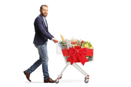 Foto de Joven caminando y empujando un carrito de compras con comida y atado con un lazo de cinta roja aislado sobre fondo blanco - Imagen libre de derechos
