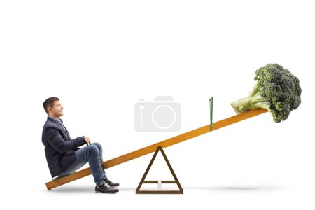 Photo for Full length profile shot of a young man and broccoli vegetable on a seesaw, healthy diet concept isolated on white background - Royalty Free Image