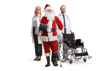 Foto de Santa Claus lesionado con un soporte para los pies y un cabestrillo para los brazos de pie con un médico y una doctora con una silla de ruedas aislada sobre fondo blanco - Imagen libre de derechos