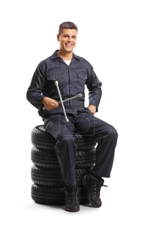 Photo for Car mechanic sitting on a pile of tires and holding a wrench tool isolated on white background - Royalty Free Image