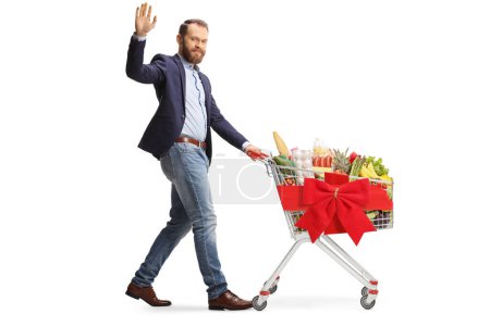 Foto de Joven barbudo en traje y jeans caminando con un carrito de compras de Navidad y saludando aislado sobre fondo blanco - Imagen libre de derechos