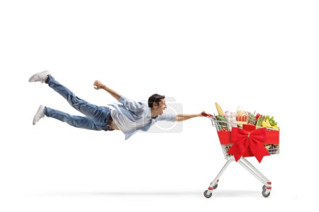 Foto de Foto completa de un joven casual volando y sosteniendo un carrito de compras con comida y lazo rojo aislado sobre fondo blanco - Imagen libre de derechos