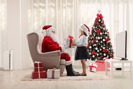 Photo for Santa claus sitting in an armchair and holding a present for a little girl in a room with a decorated tre - Royalty Free Image