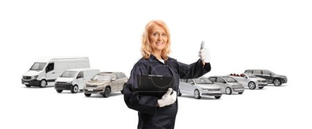 Téléchargez les photos : Mécanicien féminin en uniforme tenant un presse-papiers et gesticulant les pouces devant des véhicules isolés sur un dosseret blanc - en image libre de droit