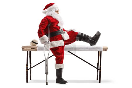 Téléchargez les photos : Prise de vue de profil du Père Noël avec un orthèse de pied et une béquille assis sur un lit médical isolé sur fond blanc - en image libre de droit