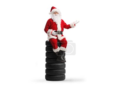 Photo for Santa claus sitting on a pile of tires and gesturing welcome sign isolated on white background - Royalty Free Image