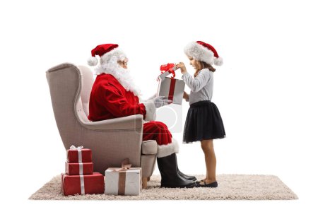 Foto de Chica impaciente abriendo una caja de regalo delante de santa claus sentado en un sillón aislado sobre fondo blanco - Imagen libre de derechos