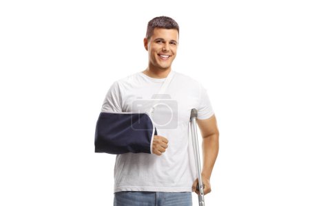 Photo for Young man with a broken arm wearing an arm splint and standing with a crutch isolated on white background - Royalty Free Image
