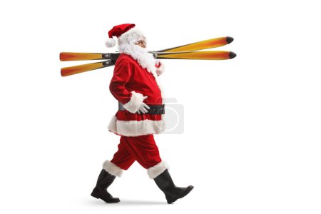 Foto de Plano de perfil de longitud completa de Santa Claus caminando y llevando esquís en el hombro aislado sobre fondo blanco - Imagen libre de derechos