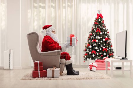 Foto de Santa Claus sentado en un sillón y sosteniendo un regalo delante de la televisión y un árbol de Navidad - Imagen libre de derechos