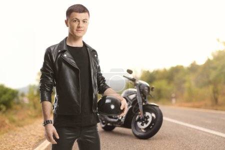 Téléchargez les photos : Jeune motard vêtu d'une veste en cuir tenant un casque de moto à côté d'un hélico sur une route - en image libre de droit