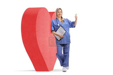 Photo for Full length portrait of a female doctor gesturing thumb up and leaning on a red heart, cardiology and health care concept - Royalty Free Image