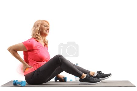Photo for Mature woman in sportswear sitting on an exercise mat and holding stiff spine isolated on white background - Royalty Free Image