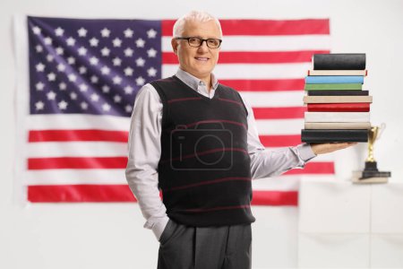 Téléchargez les photos : Professeur américain tenant une pile de livres et souriant devant un drapeau américain - en image libre de droit