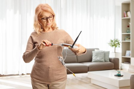 Foto de Mujer madura confundida tratando de arreglar un router con un destornillador en casa - Imagen libre de derechos