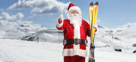Foto de Santa Claus en una montaña sosteniendo un par de esquís y señalando pulgares hacia arriba - Imagen libre de derechos
