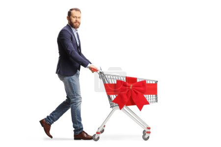 Foto de Tipo barbudo caminando y empujando y vacío carrito de compras atado con lazo de cinta roja aislado sobre fondo blanco - Imagen libre de derechos
