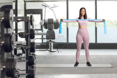 Photo for Full length portrait of a young woman in leggings and crop top exercising with an elastic rubber band at a gym - Royalty Free Image