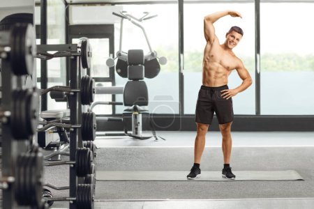 Téléchargez les photos : Portrait complet d'un homme en forme exerçant torse nu et étirant son bras à l'intérieur d'une salle de gym - en image libre de droit