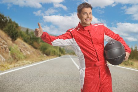 Photo for Racer in a red suit holding a helmet and hitchhiking on an open road - Royalty Free Image