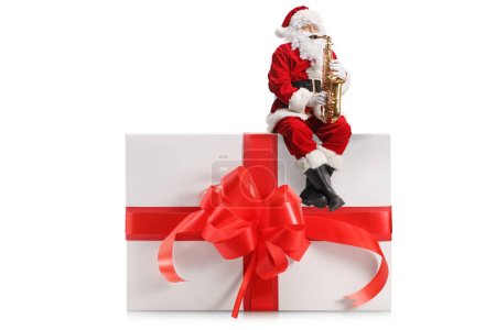 Téléchargez les photos : Père Noël jouant du saxophone et assis sur une grande boîte cadeau avec arc rouge isolé sur fond blanc - en image libre de droit
