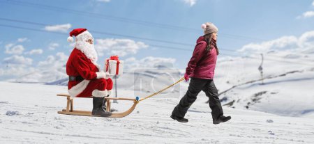 Téléchargez les photos : Fille tirant un traîneau avec le Père Noël assis et tenant un cadeau sur une colline enneigée - en image libre de droit