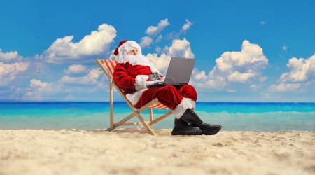 Foto de Santa Claus navegando por Internet en un ordenador portátil en la playa junto al mar - Imagen libre de derechos