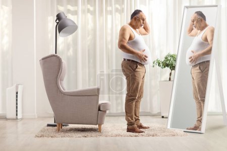 Téléchargez les photos : Malheureux mature homme tenant son gros ventre et regardant un miroir à la maison - en image libre de droit