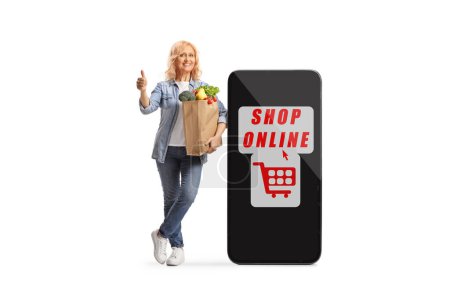 Photo for Woman with a grocery bag leaning on a big mobile phone with online shopping app and gesturing thumbs up isolated on white background - Royalty Free Image